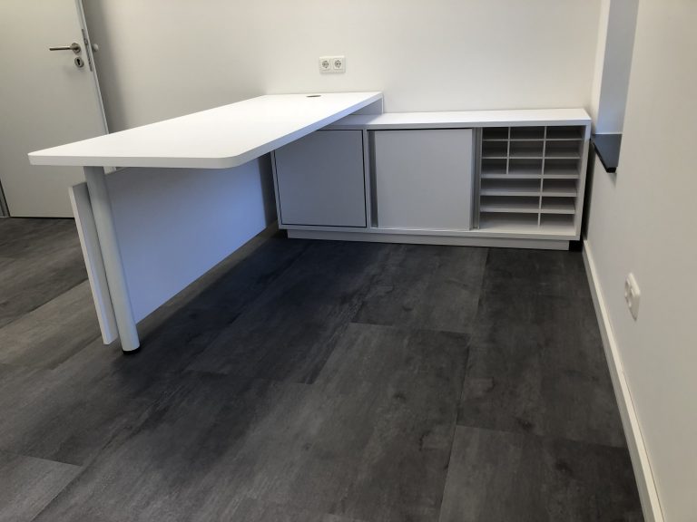 Das Bild zeigt eine moderne und funktionale Arztpraxiseinrichtung, speziell einen Schreibtisch in einem Behandlungszimmer. Der L-förmige Schreibtisch ist in mattem Weiß gehalten und passt sich perfekt der Raumgestaltung an. Er ist so konzipiert, dass er maximale Arbeitsfläche bietet, während er zugleich eine aufgeräumte Ästhetik bewahrt.
Ein besonderes Merkmal ist das integrierte Kabelmanagement: Oberhalb der Tischplatte sind Steckdosen und Anschlüsse direkt in der Wand eingelassen, und die Kabel werden unsichtbar in einem Kabelkanal verlegt. Dadurch hängen keine störenden Kabel herunter, was zur sauberen und professionellen Optik beiträgt und die Hygiene erleichtert.
Unter dem kürzeren Schenkel des Schreibtisches befindet sich ein niedriges, weißes Sideboard. Es verfügt über Schiebetüren, hinter denen sich geschlossener Stauraum verbirgt, sowie offene Fächer, die sich ideal für die Organisation von Dokumenten oder medizinischen Verbrauchsmaterialien eignen. Der Boden ist mit dunklem Laminat oder Vinyl ausgelegt und bildet einen Kontrast zu den hellen Möbeln und Wänden, was dem Raum eine zeitgemäße und klare Ausstrahlung verleiht.