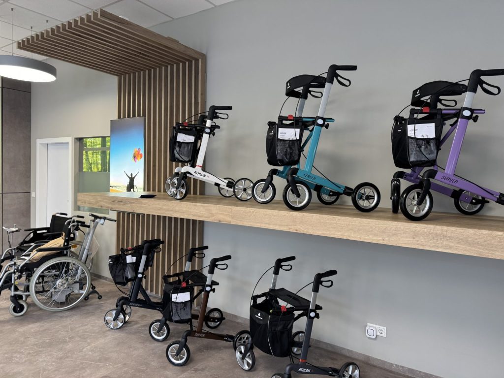 Ausstellungsraum eines Sanitätshauses für Mobilitätshilfen zeigt. Im Vordergrund sind zwei Ebenen mit verschiedenen Rollatoren zu sehen. Auf einem hellen Holzregal stehen drei Rollatoren in den Farben Weiß, Türkis und Lila. Unterhalb des Regals, auf dem Boden, sind zwei weitere Rollatoren in dunkleren Farbtönen platziert. Im Hintergrund, auf der linken Seite des Bildes, sind mehrere Rollstühle und ein silberner Rollator zu sehen. Ein kleiner Bildschirm an der Wand zeigt eine Szene mit einem geöffneten Fenster und einem Menschen, der die Arme ausstreckt, was ein Gefühl von Freiheit und Offenheit vermittelt. Die Wände des Raumes sind in einem neutralen Grau gehalten, und die Decke ist mit Holzlamellen verziert, die dem Raum eine moderne und einladende Atmosphäre verleihen