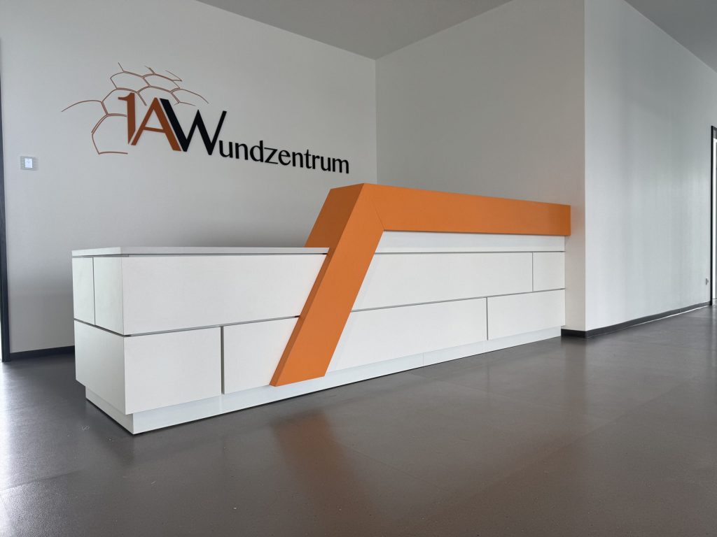 Das Bild zeigt einen modernen Empfangsbereich einer Arztpraxis, der sich durch eine markante Gestaltung auszeichnet. Im Zentrum steht eine lange, weiße Rezeptionstheke mit einer dynamischen, L-förmigen orangefarbenen Akzentleiste, die sich über die Theke zieht und ihr eine futuristische Anmutung verleiht. Die Theke selbst ist in mehrere Paneele unterteilt, die unterschiedliche Höhen und Tiefen aufweisen und so zusätzliche visuelle Textur schaffen.
An der weißen Wand hinter der Theke ist das Logo des "IAW Wundzentrum" zu sehen, mit orangefarbenen Buchstaben und einer stilisierten Waben- oder Zellstruktur, die auf den medizinischen Schwerpunkt hinweist. Der Boden ist in einem dunklen Grau gehalten und kontrastiert mit den hellen Wänden und der Rezeption. Rechts von der Theke führt ein Flur tiefer in die Praxis hinein. Die Beleuchtung ist dezent und sorgt für eine klare, aber nicht grelle Ausleuchtung des Raumes, was eine professionelle und zugleich beruhigende Atmosphäre schafft.