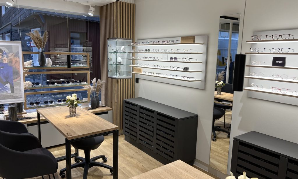 Das Bild zeigt eine moderne und stilvoll eingerichtete Verkaufsfläche, die ideal für eine Objekteinrichtung wie ein Optikergeschäft oder eine Boutique ist. Im Zentrum stehen mehrere Wandregale, auf denen zahlreiche Brillen und Sonnenbrillen ansprechend präsentiert werden. Diese Regale sind beleuchtet und in einem hellen Holzton gehalten, was die Produkte hervorhebt.
Links davon befindet sich eine Glasvitrine mit weiteren Ausstellungsstücken und daneben eine Wandverkleidung aus vertikalen Holzlamellen, die eine warme und natürliche Atmosphäre schafft. Im Vordergrund sind zwei Holztische mit schwarzen Metallgestellen zu sehen, an denen dunkle Stühle stehen. Auf einem der Tische steht eine kleine Vase mit weißen Blumen und eine Lampe, was den Beratungsbereich wohnlich gestaltet.
An der linken Seite des Raumes ist ein Schaufenster erkennbar, durch das Tageslicht einfällt und Einblicke in den Außenbereich gewährt. Ein großes Werbebanner mit einer Person in blauer Kleidung ist ebenfalls zu sehen. Rechts im Bild befindet sich ein großer Spiegel, der den Raum optisch vergrößert und zusätzliche Brillenpräsentationen auf einem weiteren Regal daneben reflektiert. Unterhalb der Brillenpräsentationen stehen dunkle Sideboards mit Lamellentüren, die als Stauraum dienen. Der Boden ist mit hellem Holzparkett oder Laminat ausgelegt, was die helle und einladende Gesamtwirkung der Objekteinrichtung unterstreicht.