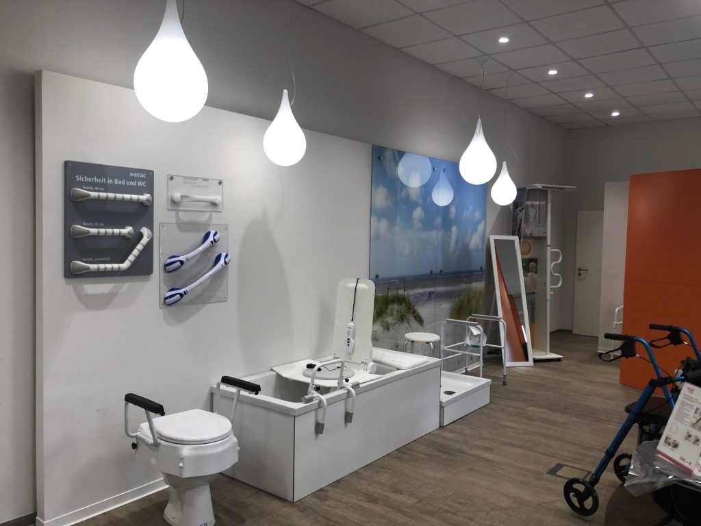 Das Bild zeigt einen modern und hell gestalteten Ausstellungsraum eines Sanitätshauses, der verschiedene Produkte für die Bad- und WC-Sicherheit sowie Mobilitätshilfen präsentiert – ein hervorragendes Beispiel für eine funktionale Sanitätshauseinrichtung. An der linken weißen Wand sind verschiedene Haltegriffe und Stützen für Bad und WC auf Tafeln montiert, die die "Sicherheit in Bad und WC" thematisieren.
Im mittleren Bereich des Raumes steht ein komplett ausgestattetes Badewannen-System mit einem integrierten Badewannenlift, das die Zugänglichkeit und Nutzung für Menschen mit Mobilitätseinschränkungen erleichtert. Daneben ist eine rollstuhlgerechte Duschwanne oder ein ähnliches Einstiegssystem zu sehen. Davor steht eine erhöhte Toilettensitzerhöhung mit Armlehnen.
Eine großflächige Wandgrafik im Hintergrund zeigt eine ruhige Strandlandschaft mit Himmel und Meer, was dem Raum eine angenehme Atmosphäre verleiht. Die Deckenbeleuchtung besteht aus mehreren modernen, tropfenförmigen Pendelleuchten, die ein warmes, diffuses Licht spenden.
Rechts im Bild sind weitere Mobilitätshilfen erkennbar, darunter ein blauer Rollator. Ein großer Spiegel und ein Duschhocker ergänzen die Ausstattung. Der Boden ist mit einem warmen Holz- oder Laminatboden ausgelegt, der zum einladenden Gesamtambiente dieser Sanitätshauseinrichtung beiträgt.