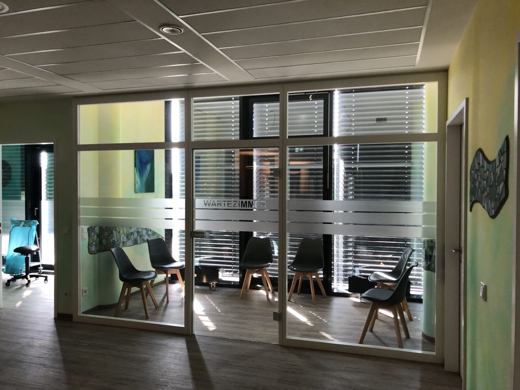 Das Bild zeigt den modernen Wartebereich einer Arztpraxis, dessen Einrichtung auf Komfort und Funktionalität ausgelegt ist. Eine große Glaswand mit der Aufschrift "WARTEZIMMER" trennt den Hauptwartebereich von einem angrenzenden Flur oder einem weiteren Raum. Die Glasflächen sind teilweise satiniert, um etwas Privatsphäre zu bieten.
Im Wartezimmer selbst stehen mehrere dunkelgraue Stühle mit Holzbeinen, die eine einladende Sitzgelegenheit bieten. Durch die großen Fenster hinter den Stühlen fällt Sonnenlicht in den Raum, was für eine helle und freundliche Atmosphäre sorgt. Die Fenster sind mit Jalousien versehen, um die Lichtverhältnisse nach Bedarf regulieren zu können.
Die Wände im Flurbereich sind in einem beruhigenden Hellgrün und Gelb gestrichen. An der rechten Wand hängt ein dekoratives, wellenförmiges Kunstobjekt in Blau und Grau. Links im Bild ist ein weiterer Bereich mit einem türkisfarbenen Stuhl und einem kleinen Tisch erkennbar, was auf einen separaten Beratungs- oder Untersuchungsraum hindeutet. Der Boden besteht aus hellem Holz- oder Laminat und trägt zur warmen und freundlichen Gesamterscheinung der Arztpraxiseinrichtung bei.