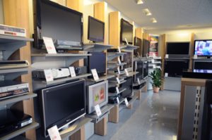 Das Bild zeigt das Innere eines Elektro- und Elektronikgeschäfts, das sich auf die Präsentation von Fernsehgeräten und Unterhaltungselektronik spezialisiert hat. Entlang der Wände sind Reihen von Fernsehern unterschiedlicher Größen und Modelle auf Regalsystemen montiert oder auf Konsolen platziert. Die Geräte sind eingeschaltet und zeigen verschiedene Bilder oder Videos, was die Funktionalität und Bildqualität demonstriert.
Die Regalsysteme bestehen aus hellgrauen Metallregalböden und einer hellen Holzverkleidung, die eine moderne und übersichtliche Präsentation ermöglicht. Unter einigen Fernsehern sind zusätzliche Geräte wie DVD-Player, Receiver oder Soundbars auf den Regalböden ausgestellt. Der Gang zwischen den Reihen ist breit genug, um Kunden eine gute Sicht auf die Produkte zu ermöglichen.
Im Hintergrund sind weitere Ausstellungsbereiche zu erkennen, und der Boden ist in einem neutralen Farbton gehalten, der die Aufmerksamkeit auf die ausgestellten Elektronikartikel lenkt. Ein Topf mit einer grünen Pflanze ist als dezenter Dekorationsakzent in einem der Gänge platziert. Die gesamte Einrichtung ist darauf ausgelegt, eine große Auswahl an Elektronikprodukten ansprechend und funktional zu präsentieren.